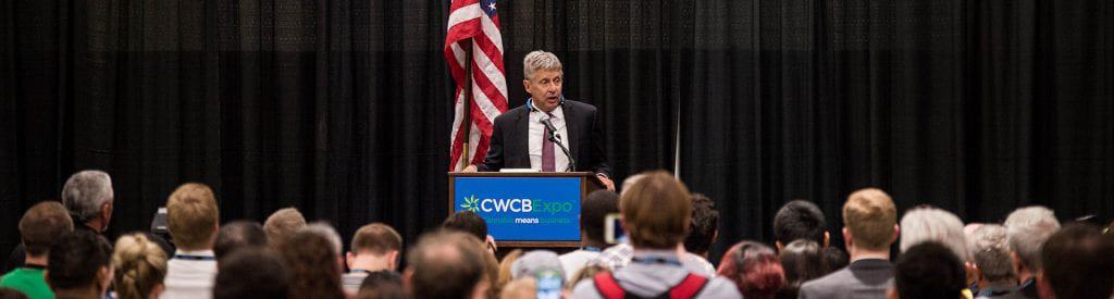 A man speaks at a podium in front of an audience at the CWCB Expo in Los Angeles. The podium, adorned with the CWCB Expo logo and site information, stands before an attentive crowd. An American flag waves in the background as he delves into topics from the Cannabis Legal Workshop hosted by Cal NORML. CA Norml