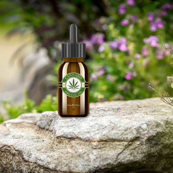 A brown glass dropper bottle labeled "Cannabis Organic Oil" sits on a large rock. The background features a blurred mix of greenery and pink flowers, suggesting a natural, outdoor setting. CA Norml