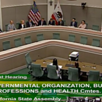 A governmental meeting room in Sacramento with green chairs and a U-shaped arrangement of desks. Several people are seated while a few stand or sit at a central desk. American and California flags are visible. Text on the screen mentions a joint hearing of various CA Committees about vaping regulations. CA Norml