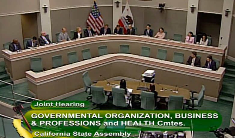 A governmental meeting room in Sacramento with green chairs and a U-shaped arrangement of desks. Several people are seated while a few stand or sit at a central desk. American and California flags are visible. Text on the screen mentions a joint hearing of various CA Committees about vaping regulations. CA Norml