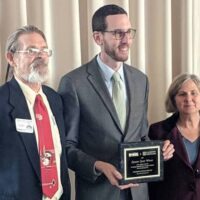 Three people stand in a row, smiling. The man in the center, wearing glasses and a suit, holds a plaque. The man on the left has glasses, a beard, and wears a suit with a red tie. The woman on the right wears a dark jacket. A beige curtain is in the background. CA Norml