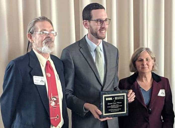 Three people stand in a row, smiling. The man in the center, wearing glasses and a suit, holds a plaque. The man on the left has glasses, a beard, and wears a suit with a red tie. The woman on the right wears a dark jacket. A beige curtain is in the background. CA Norml