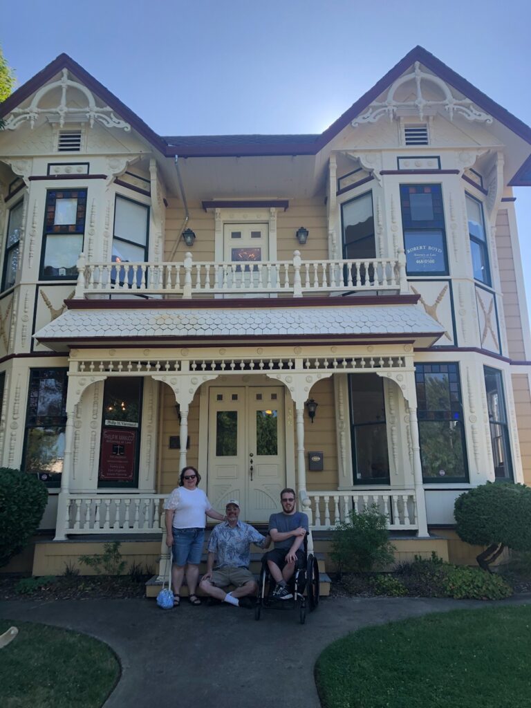 A woman and two men, with one of the men in a wheelchair, pose for a photo in front of a large, ornate yellow house with a covered porch and intricate woodwork. They appear to be enjoying a sunny day, standing on a concrete path flanked by green shrubs. CA Norml