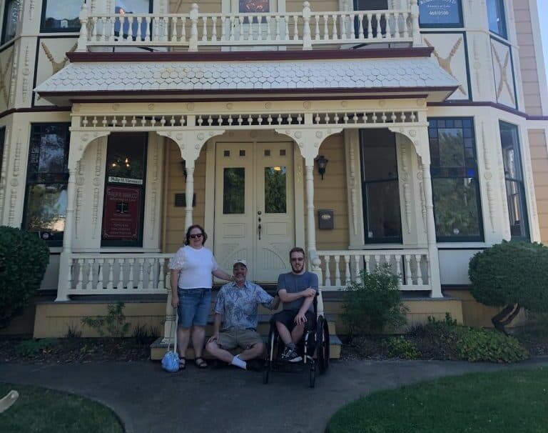 A woman and two men, with one of the men in a wheelchair, pose for a photo in front of a large, ornate yellow house with a covered porch and intricate woodwork. They appear to be enjoying a sunny day, standing on a concrete path flanked by green shrubs. CA Norml