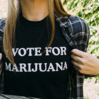 A person with long hair wearing a black t-shirt with the text "VOTE FOR MARIJUANA" printed on it holds open their plaid shirt to display the message. Against a backdrop of green foliage and sunlight, they emphasize support for CA Special Elections on May 12. CA Norml