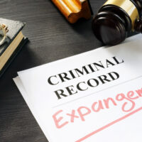 A document labeled "CRIMINAL RECORD" with the word "Expungement" written in red sits on a dark desk. Nearby are a pair of glasses, a closed book, and a judge's gavel. The scene signifies steps taken in California for clearing past marijuana crimes from individual records. CA Norml