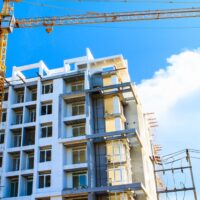 A modern, multi-story building under construction with exposed concrete and scaffolding is seen, set against a vibrant blue sky with a few clouds in West Hollywood. A tall yellow crane signals active construction work, underscoring the city's growth amid its progressive initiatives like the smoking ban. CA Norml