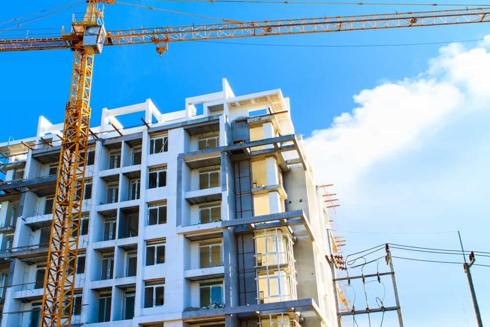 A modern, multi-story building under construction with exposed concrete and scaffolding is seen, set against a vibrant blue sky with a few clouds in West Hollywood. A tall yellow crane signals active construction work, underscoring the city's growth amid its progressive initiatives like the smoking ban. CA Norml