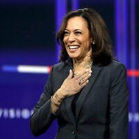 A woman in a dark blazer stands on stage, smiling and holding her hand to her chest. The background features blurred, dark blue elements and indistinct text. She appears to be speaking or acknowledging an audience, reminiscent of Kamala Harris when she was introduced as Biden's running mate. CA Norml