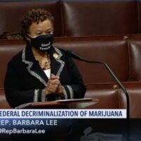 A person stands at a podium in a legislative chamber, wearing a black mask with white text that may read "Good Trouble." The screen shows text: "Federal Decriminalization of Marijuana," "Rep. Barbara Lee," and her handle @RepBarbaraLee. The C-SPAN logo is visible. California NORML supports this 2021 marijuana reform effort. CA Norml