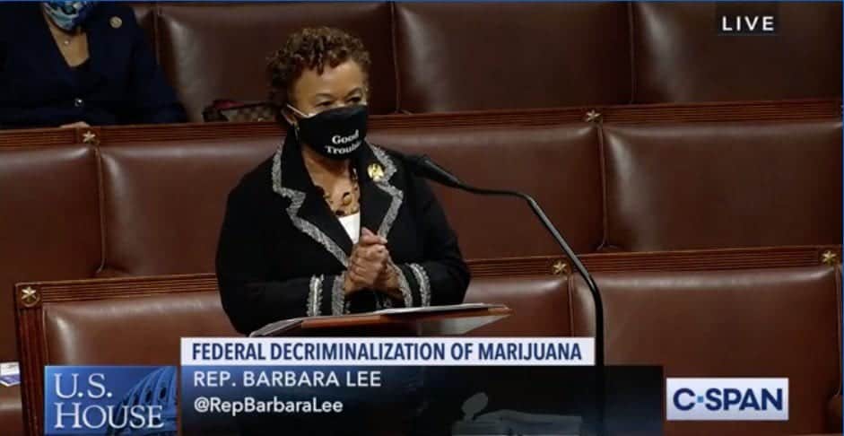 A person stands at a podium in a legislative chamber, wearing a black mask with white text that may read "Good Trouble." The screen shows text: "Federal Decriminalization of Marijuana," "Rep. Barbara Lee," and her handle @RepBarbaraLee. The C-SPAN logo is visible. California NORML supports this 2021 marijuana reform effort. CA Norml