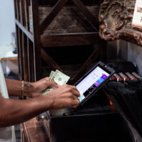 A person is standing at a cash register, holding a few dollar bills in one hand and operating a touch screen with the other. In the background, there is a sign for a loyalty points program and a shelf with various items promoting local businesses. CA Norml