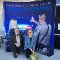 A man and a woman smile for a photo in front of a California Highway Patrol banner promoting impaired driving awareness. The banner, resembling a scene from a CHP Report, shows a patrol officer administering a sobriety test to a woman. CA Norml