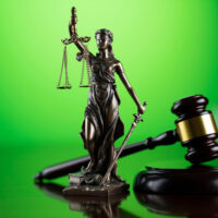 A bronze statue of Lady Justice, holding scales and a sword, stands in front of a green background. A wooden judge's gavel and sound block are placed beside the statue, symbolizing law and justice. The scene commemorates the Legal Seminar held in June 2021 by California NORML. CA Norml