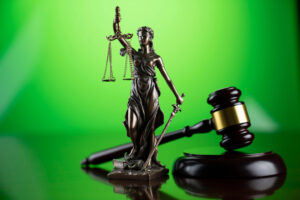 A bronze statue of Lady Justice, holding scales and a sword, stands in front of a green background. A wooden judge's gavel and sound block are placed beside the statue, symbolizing law and justice. The scene commemorates the Legal Seminar held in June 2021 by California NORML. CA Norml