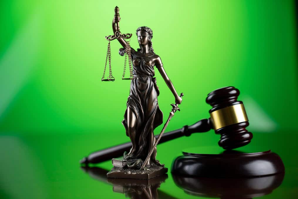 A bronze statue of Lady Justice, holding scales and a sword, stands in front of a green background. A wooden judge's gavel and sound block are placed beside the statue, symbolizing law and justice. The scene commemorates the Legal Seminar held in June 2021 by California NORML. CA Norml