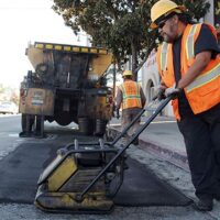 A CA state employee in an orange safety vest operates a compactor to pave over a section of road. Another worker in the background directs traffic while a dump truck is parked nearby. A traffic cone and a convenience store can be seen in the vicinity. CA Norml