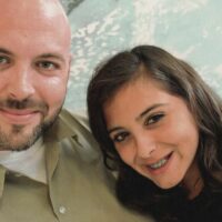 A man with a shaved head and a beard is smiling at the camera. Next to him, a young woman with long, dark hair and braces is also smiling. They appear to be taking a selfie against a background featuring abstract, natural patterns. This moment captures Luke Scarmazzo, a former Federal Medical Marijuana Prisoner from California, enjoying life after his release. CA Norml