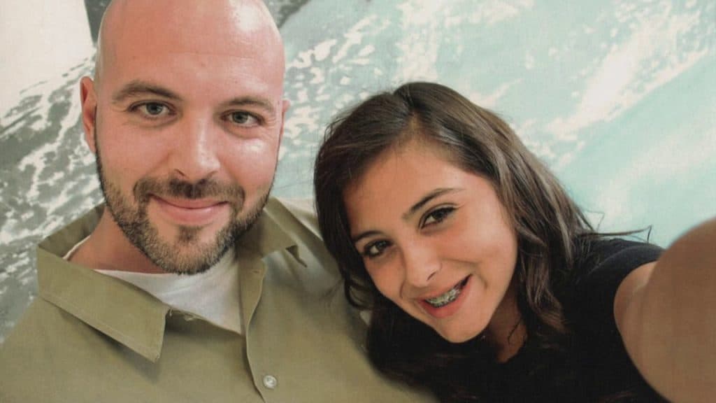 A man with a shaved head and a beard is smiling at the camera. Next to him, a young woman with long, dark hair and braces is also smiling. They appear to be taking a selfie against a background featuring abstract, natural patterns. This moment captures Luke Scarmazzo, a former Federal Medical Marijuana Prisoner from California, enjoying life after his release. CA Norml