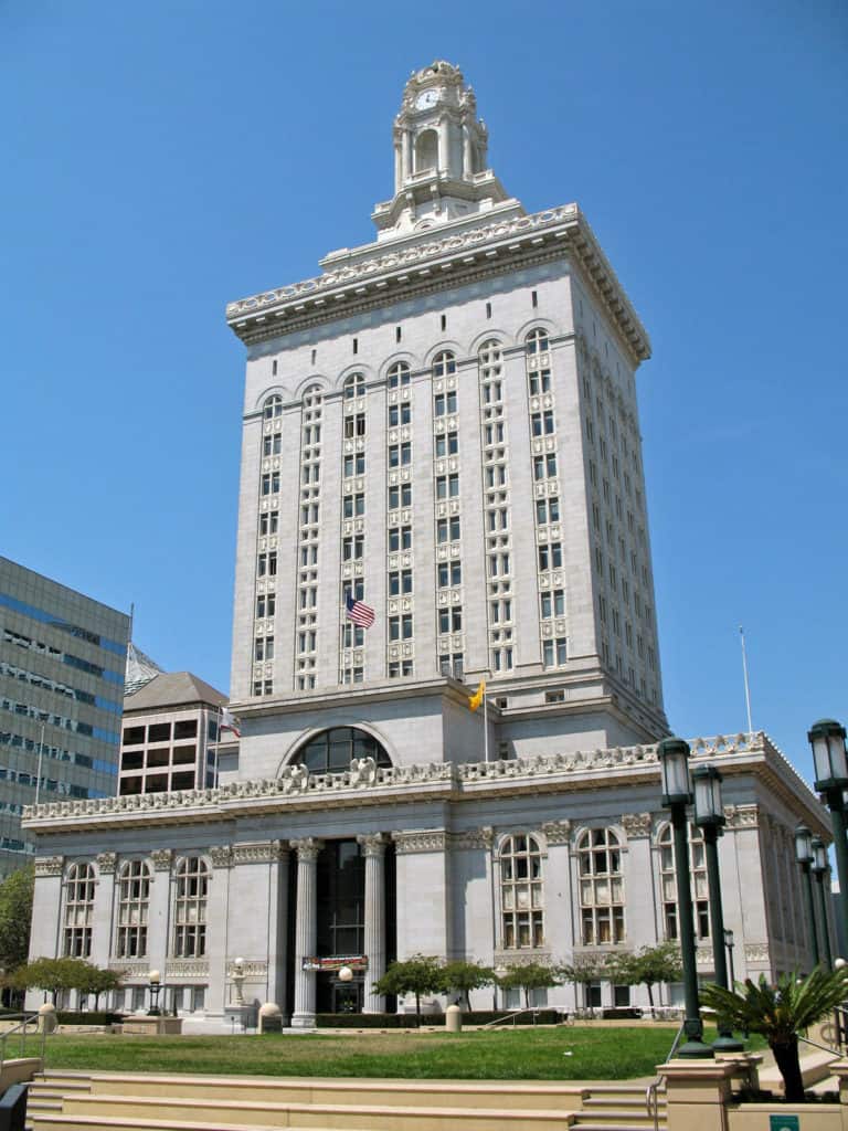 A tall, historic building with a clock tower stands under a clear blue sky. The structure features an ornate design with large windows and decorative columns. Two flags, one American and one unknown, are displayed near the entrance. Surrounding buildings and a small park area with lampposts and trees are visible in the foreground. CA Norml