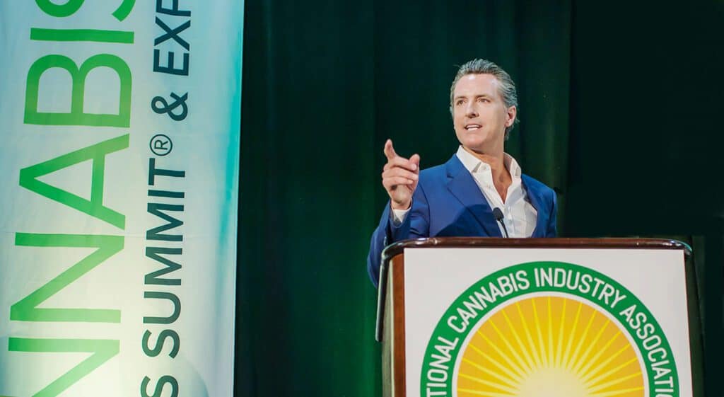 A person in a blue suit speaks at a podium adorned with the National Cannabis Industry Association logo. Behind them is a vertical banner that reads "Cannabis Business Summit & Expo." The dark background underscores the importance of issues like the Newsom recall, urging attendees to vote no. CA Norml