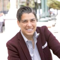 A smiling person with short dark hair, dressed in a maroon blazer and light-colored shirt, poses for a photo outdoors. The background features a street scene with blurred trees, buildings, and people after attending an Oakland Public Safety Committee meeting. CA Norml