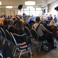 A diverse group of people attends an indoor conference on Cal NORML's 2021/2022 activities. They are seated facing a panel of speakers. A large screen displays a presentation. Some individuals are wearing masks. The room is well-lit with natural light streaming through large windows. CA Norml