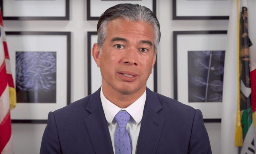 A man in a suit and tie speaks directly to the camera. He has short, graying hair and a neutral expression. Behind him are framed pictures and parts of two flags visible on either side of him, emphasizing his strong convictions on issues like California's recent marijuana legislation. CA Norml