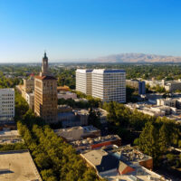 Aerial view of a cityscape with a mix of historic and modern buildings, including a prominent tall brick building with a spire, set against a backdrop of leafy greenery and distant mountains under a clear blue sky. CA Norml