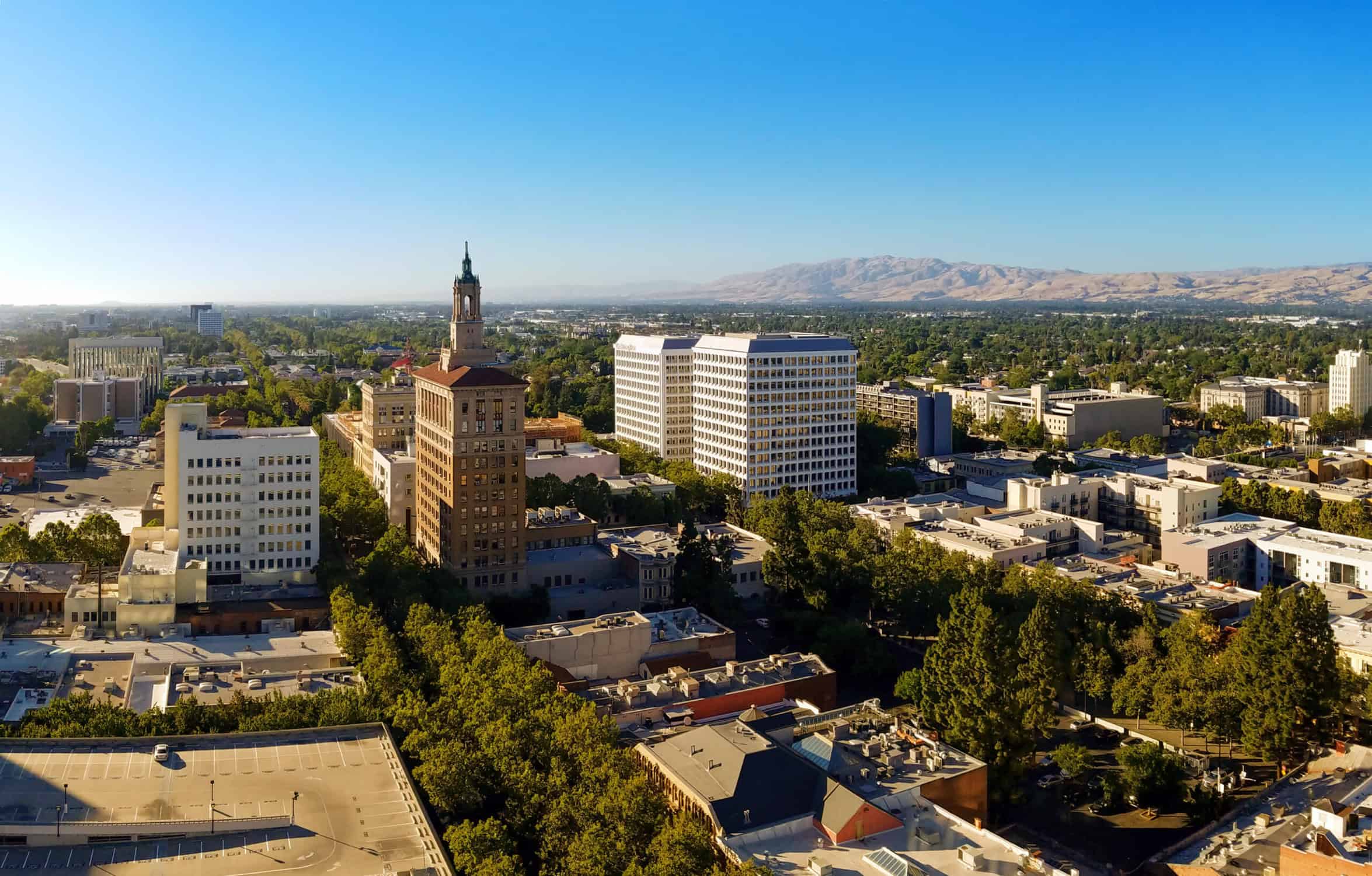 Aerial view of a cityscape with a mix of historic and modern buildings, including a prominent tall brick building with a spire, set against a backdrop of leafy greenery and distant mountains under a clear blue sky. CA Norml