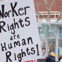 A person holds a large sign at a rally or protest. The sign reads "Worker Rights are Human Rights!" In the background, there are other people and an urban setting with buildings and leafless trees as Californians gather to advocate for employment rights. CA Norml