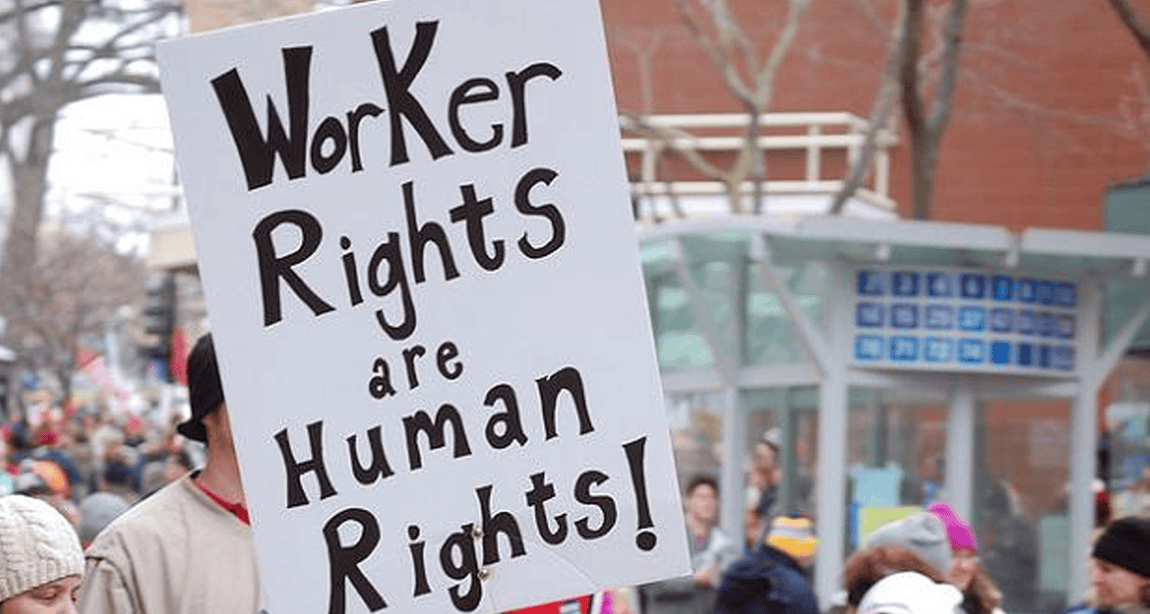 A person holds a large sign at a rally or protest. The sign reads "Worker Rights are Human Rights!" In the background, there are other people and an urban setting with buildings and leafless trees as Californians gather to advocate for employment rights. CA Norml