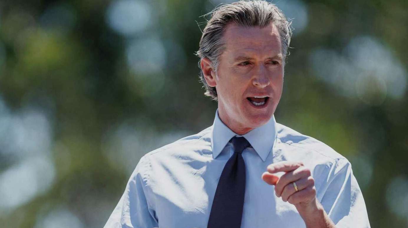 A man with gray hair, wearing a light blue dress shirt and dark tie, is speaking passionately outdoors about employment rights. His mouth is open and he is pointing with his right hand. The background is blurred and appears to be greenery. CA Norml