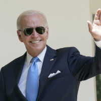 A smiling man wearing a dark suit, light blue tie, and sunglasses is waving with his right hand. He is outside, with a white wall and a window in the background, reminiscent of scenes from President Biden's recent public appearances. CA Norml