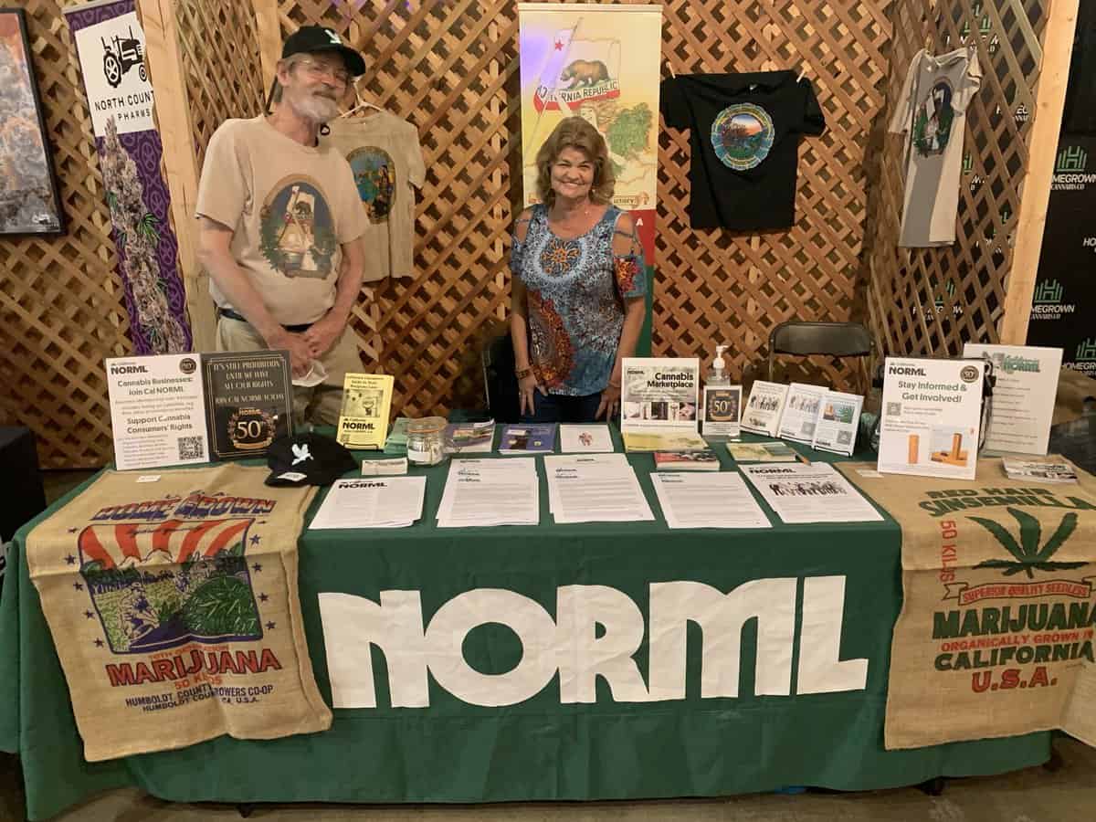 Two individuals stand behind a booth with a table covered by a cloth displaying "Cal NORML." The table is adorned with pamphlets, brochures, and other literature about marijuana legalization, including 2022 accomplishments and plans for 2023. The background features lattice panels with hanging T-shirts and banners. CA Norml