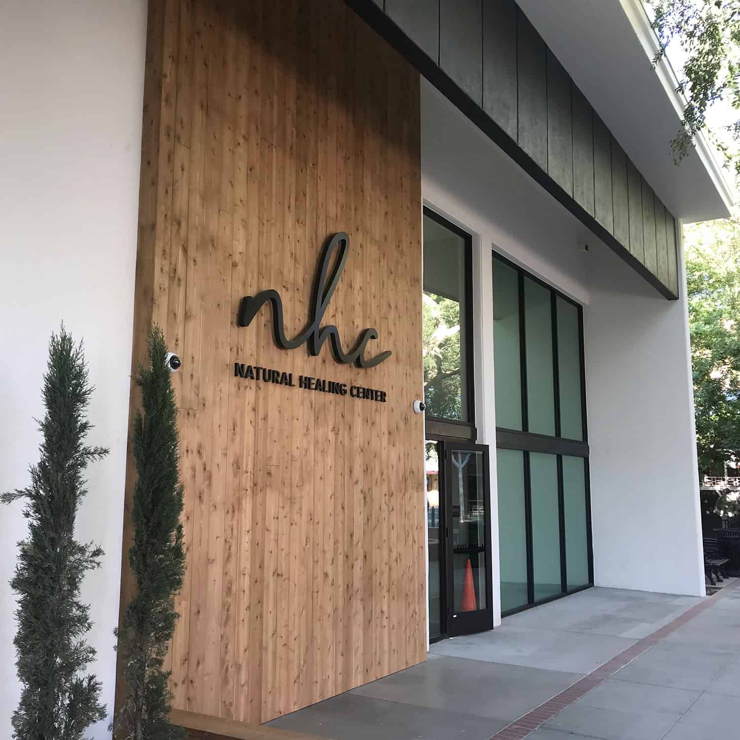 The photo shows the exterior of a building with large glass windows and a wooden facade. The sign on the wood reads "NHC Natural Healing Center" in black letters. There are two tall, narrow trees in front of the dispensary. CA Norml