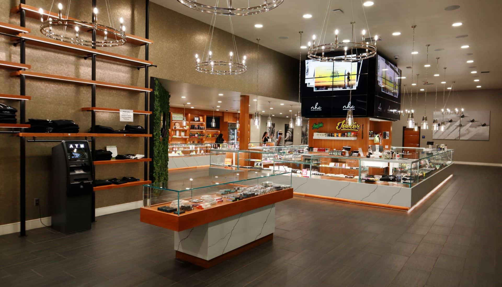 A modern dispensary interior, also known as a Healing Center, features glass display cases filled with products, sleek wooden shelving on the left wall, and a well-lit ceiling with contemporary light fixtures. The room has a clean, organized appearance blending natural healing elements with modern design. CA Norml