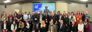 A large group of people posing for a group photo in a conference room. Behind them, a projected slide reads "California Citizen Lobby Day in Sacramento 5/8/2023," along with other text. The group appears to be at an event or meeting, with a diverse range of attendees smiling at the camera. CA Norml