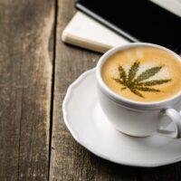 A cup of coffee sits on a white saucer atop a rustic wooden table. The frothy top of the coffee features a leaf design created with green powder, reminiscent of some cannabis lounges. In the background, there is a partially opened notebook and a black smartphone. CA Norml