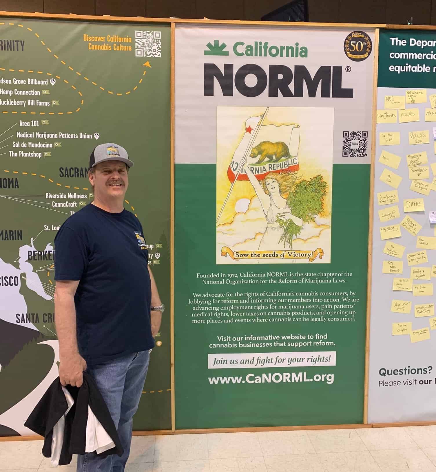 A man stands smiling next to a large California NORML poster at an event on July 25, 2023. The poster features a woman holding a flag and a marijuana leaf, offering information about Cal NORML’s advocacy for marijuana law reform. The man is wearing a cap and holding a bag. CA Norml