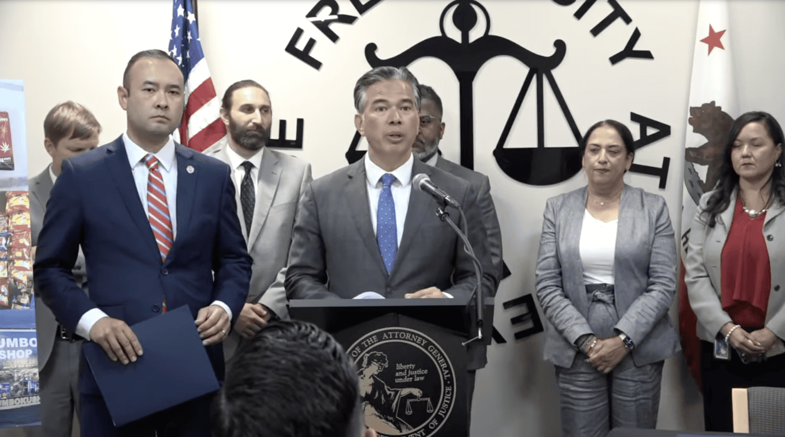 A group of seven individuals stands in front of a "Franchise Tax Board" wall display. The central figure, AG Bonta, speaking at a podium with a seal, is flanked by a diverse group of people in formal attire. An American flag and the California state flag are visible in the background. CA Norml