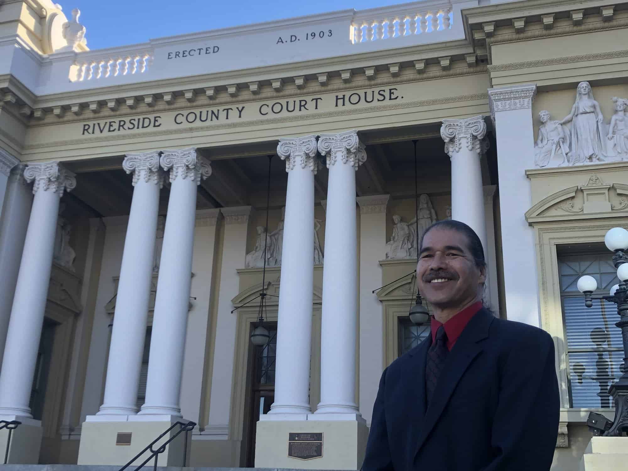 A man in a dark suit and red shirt stands smiling in front of the historic Riverside County Courthouse. The building features grand white columns, intricate stone sculptures, and a sign that reads "Riverside County Courthouse." The date "A.D. 1903" is visible. This is James M. Taylor, Criminal Defense Attorney. CA Norml