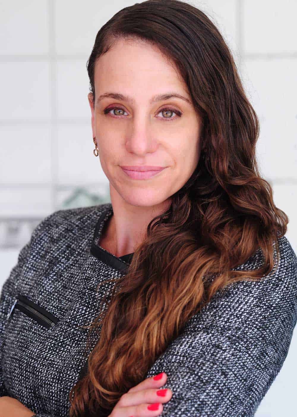 A woman with long, wavy brown hair, standing against a white-tiled wall, looks directly at the camera with a slight smile. Wearing a gray textured top with black trim, Jennie Stepanian has her arms crossed. She has light eye makeup, wears small earrings, and her nails are painted red. CA Norml