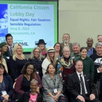 A large, diverse group of people posing for a photo in a conference room. A presentation screen behind them reads "California Citizen Lobby Day: Equal Rights, Fair Taxation, Sensible Regulation, May 4, 2023, Sacramento, CA." Everyone is smiling and celebrating the event and their 2023 accomplishments with Cal NORML. CA Norml