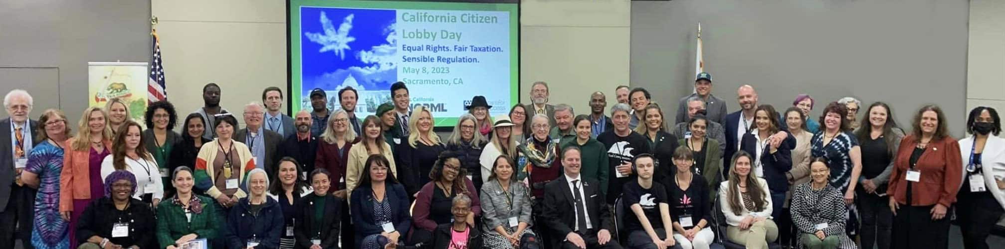 A large, diverse group of people posing for a photo in a conference room. A presentation screen behind them reads "California Citizen Lobby Day: Equal Rights, Fair Taxation, Sensible Regulation, May 4, 2023, Sacramento, CA." Everyone is smiling and celebrating the event and their 2023 accomplishments with Cal NORML. CA Norml