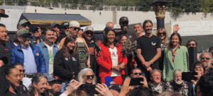 A diverse group of people, including Mayor Breed, smile and pose for a photo outdoors. Some hold a large belt that appears to be an award. The backdrop showcases greenery, a white wall, and a small structure, all under the sunny skies of SF Weed Week. CA Norml