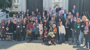 A diverse group of people poses together outside a large building with double doors and steps in Sacramento. Many wear conference badges, indicating it's Lobby Day on May 6. Some are seated, while others stand, representing mixed ages and backgrounds. Everyone is smiling and facing the camera. CA Norml