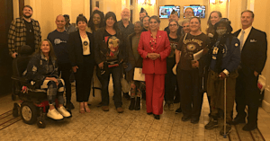 A diverse group of people stand in a lobby, some holding papers. At the center, a woman in a red pantsuit smiles. One person uses a wheelchair. The environment appears formal, with chandeliers and carpeted flooring, suggesting an official or celebratory event perhaps related to Compassion Programs. CA Norml