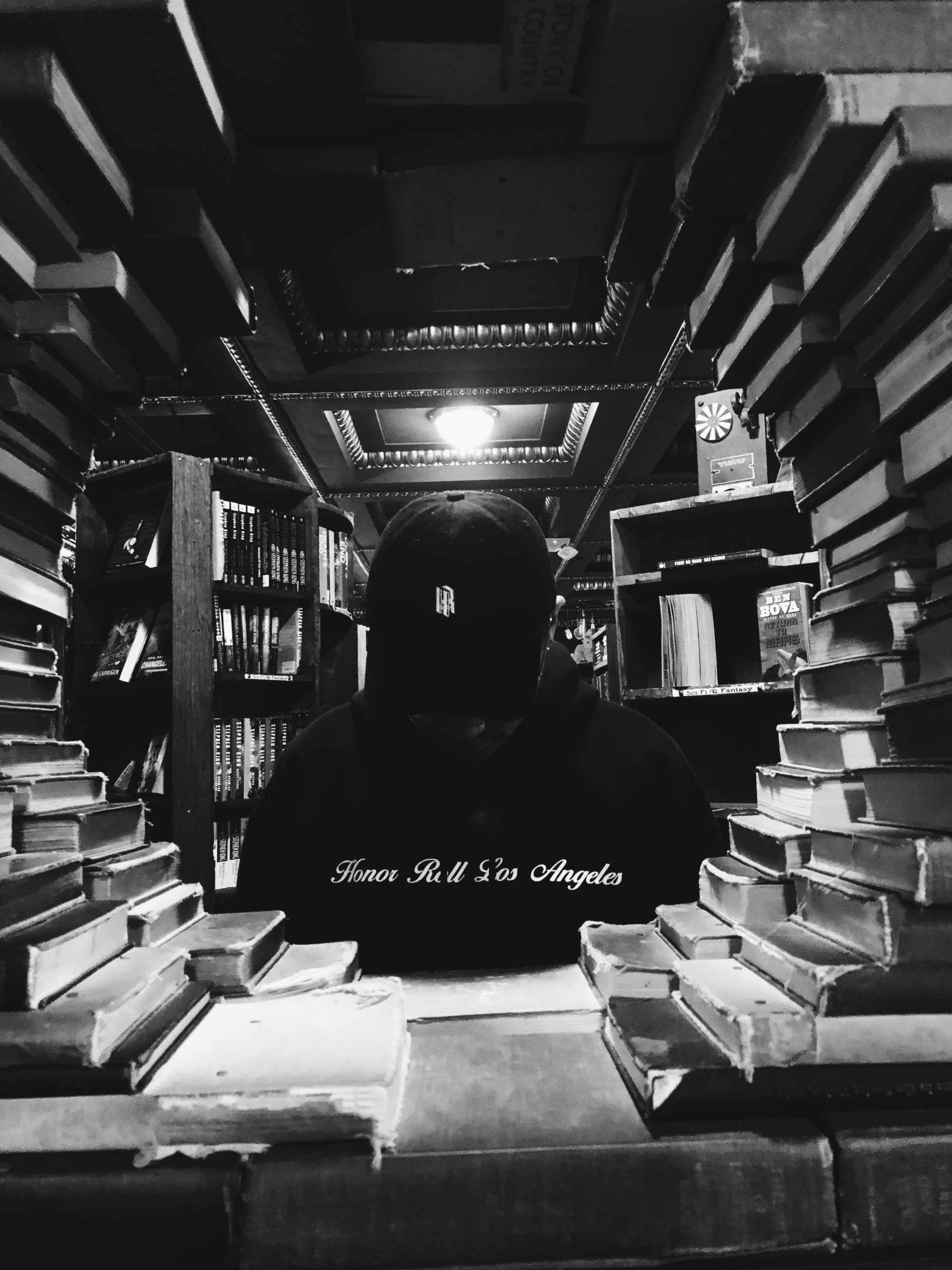 A person wearing a cap and hoodie stands in a dimly lit library, seen through a tunnel of stacked books. Shelves of books and ornate ceiling details are visible in the background, creating a cozy and mysterious atmosphere, reminiscent of an Honor Roll Delivery. The image is in black and white. CA Norml