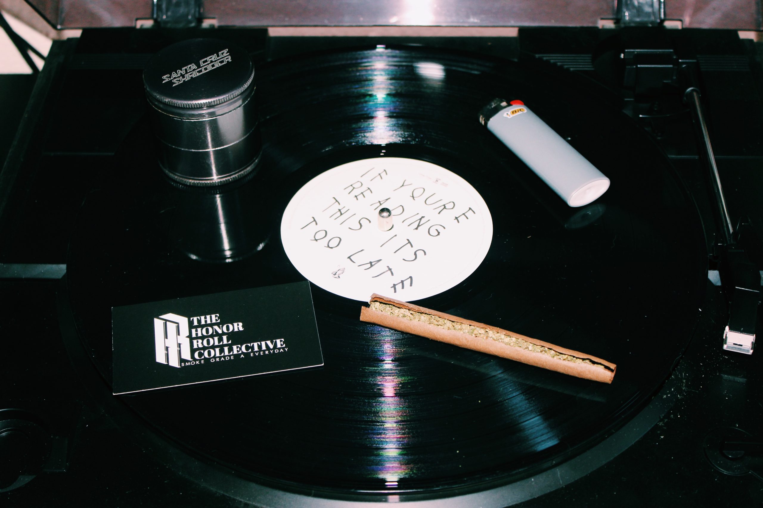 A close-up of a black vinyl record displayed on a turntable. Items on the record include a gray lighter, a rolled-up lighter tobacco pipe, a small round container, and a black business card labeled "The Honor Roll Collective." The record label reads "If You're Reading This, It's Too Late," courtesy of Honor Roll Delivery. CA Norml